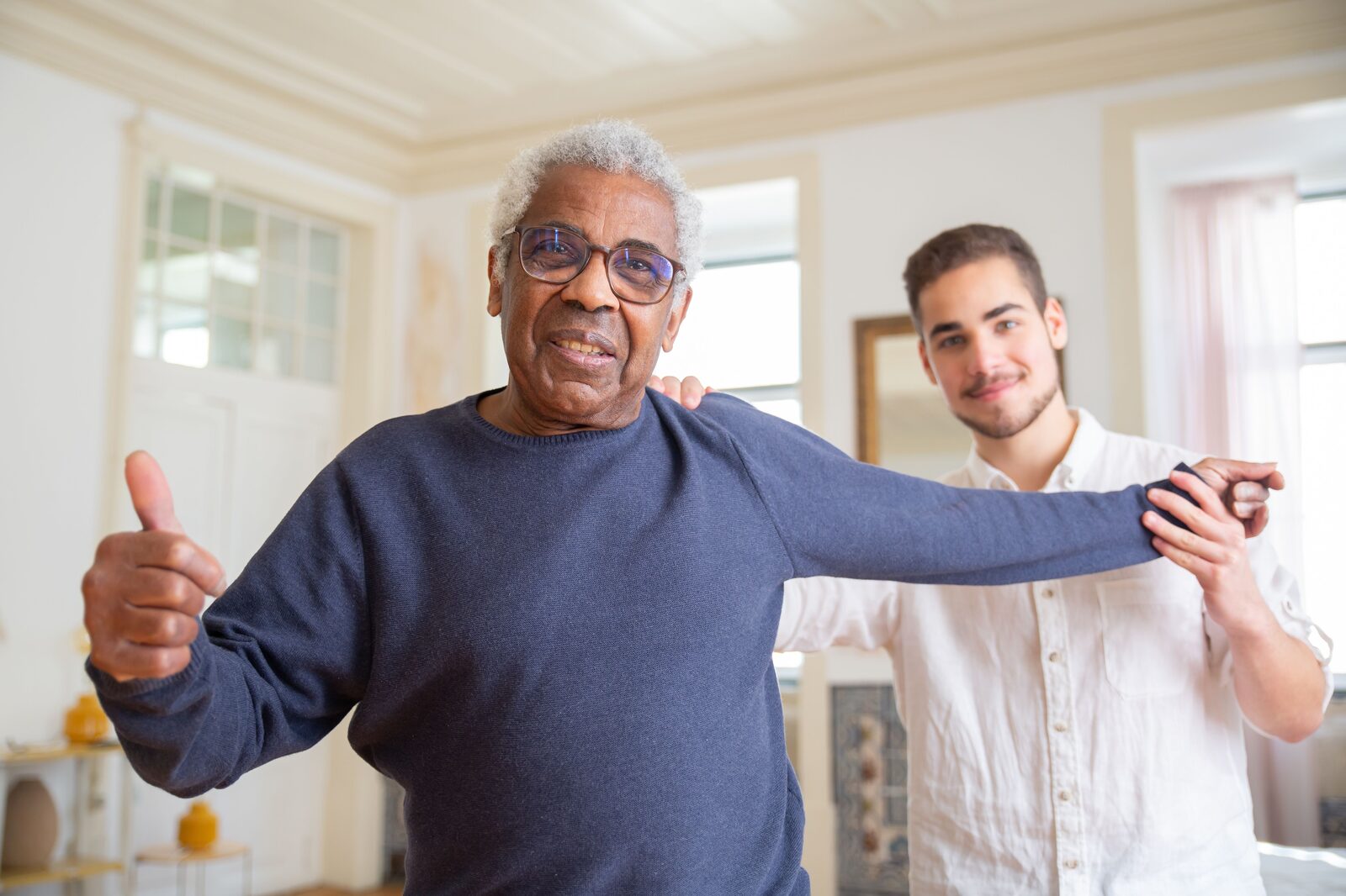 A younger man supporting an older man's arm as they walk through a bright living room
