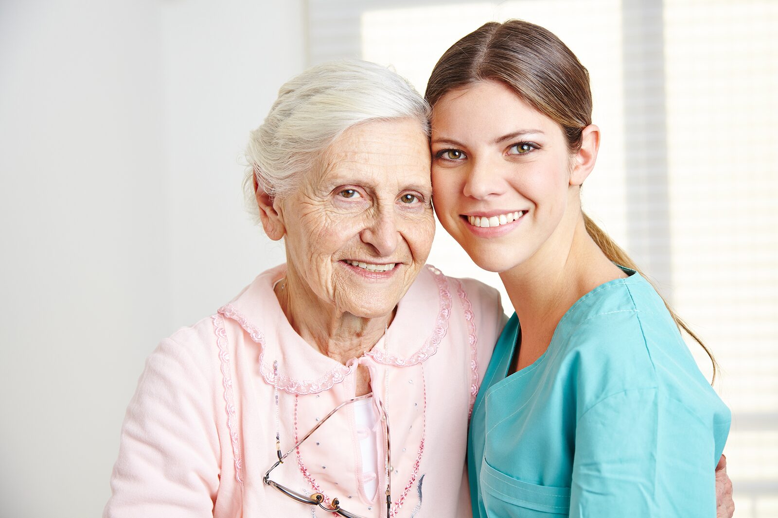 A smiling caregiver with her arm around an older woman in a bright room at home