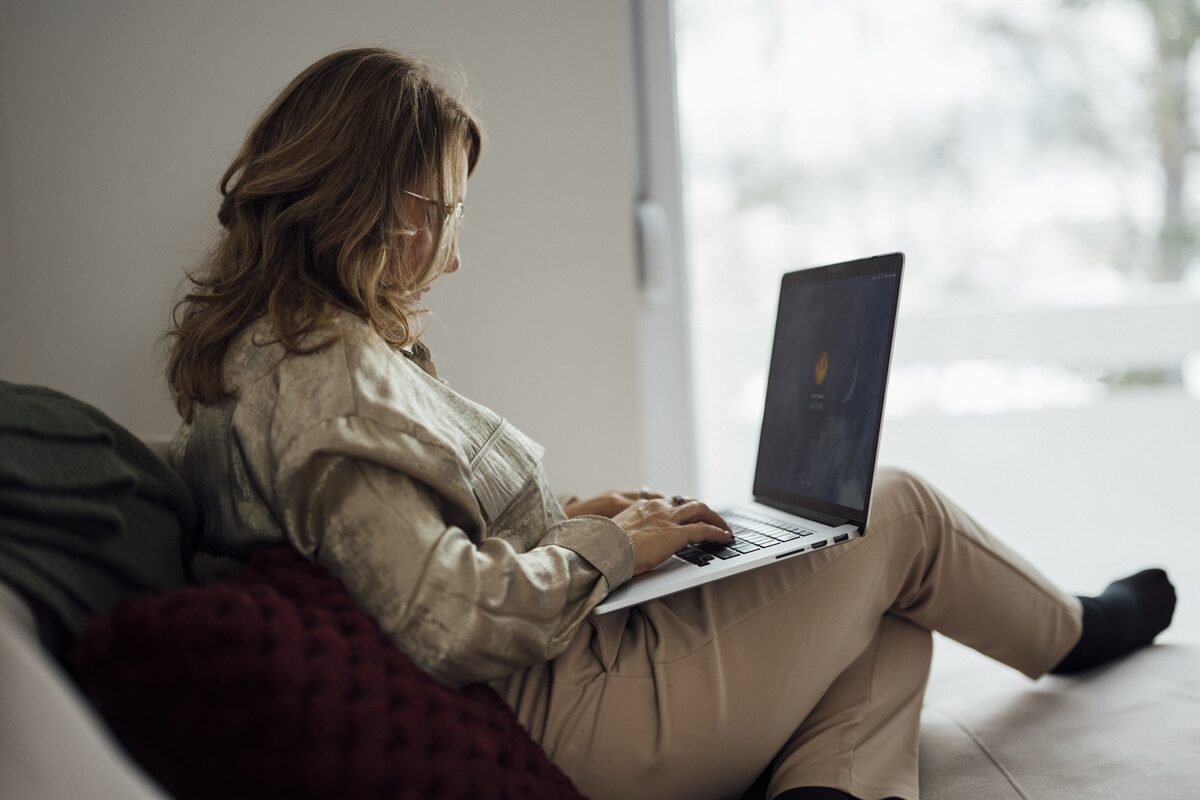 A woman on a sofa at home reviewing caregiver profiles on a laptop